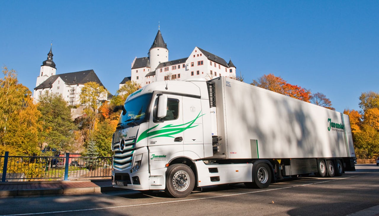 Weißer Mercedes-LKW mit grünem Firmenlogo vor einem Schloss und bunten Herbstbäumen.